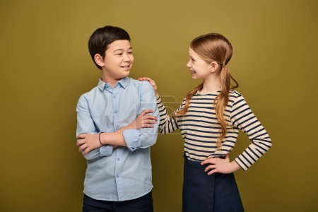Lächelndes rothaariges Preteen-Mädchen, das sich umarmt und asiatische Freundin ansieht, die Hände faltet, während internationaler Kinderschutztag auf khakifarbenem Hintergrund