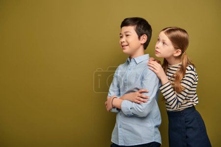 Photo for Redhead girl in striped blouse hugging smiling asian friend crossing and and looking away during child protection day celebration on khaki background with copy space - Royalty Free Image
