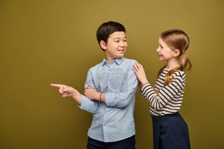Smiling redhead girl in striped blouse hugging preteen asian friend pointing with finger away while showing something during child protection day celebration on khaki background