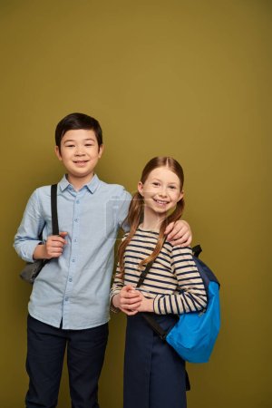 Lächelnder asiatischer Junge mit Rucksack umarmt rothaarige Freundin und blickt während der Feier zum internationalen Kinderschutztag auf khakifarbenem Hintergrund in die Kamera