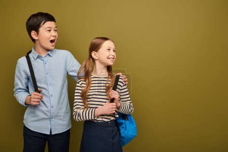 Excited asian boy with opened mouth with backpack opening mouth and hugging cheerful redhead friend and looking away during child protection day celebration on khaki background