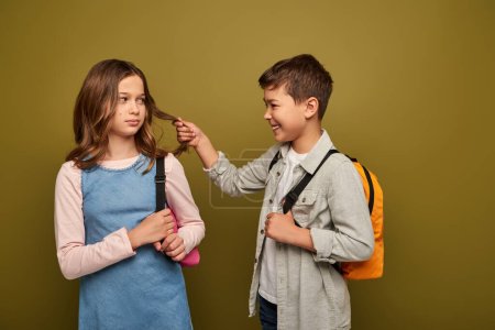 Smiling multiracial boy with backpack pulling hair of preteen girl while getting attention during international child protection day celebration on khaki background