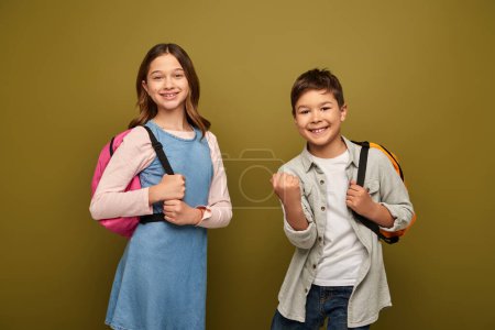 Excited multiracial boy with backpack showing yes gesture and looking at camera near friend in dress during child protection day celebration on khaki background