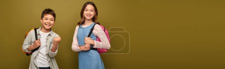 Carefree multiracial boy with backpack showing yes gesture at camera near friend during child protection day celebration on khaki background with copy space, banner 