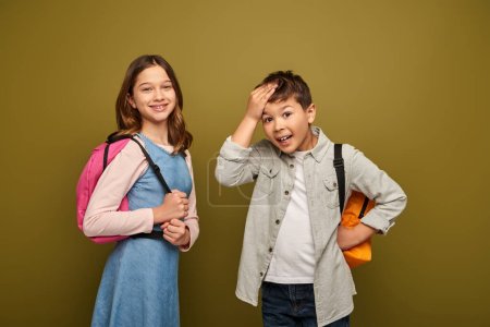 Erstaunter multirassischer Junge berührt Kopf und blickt in Kamera neben lächelndem Freund mit Rucksack während globalem Kinderschutztag auf khakifarbenem Hintergrund