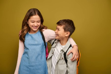 Carefree preteen and multiethnic kids in casual clothes with backpacks hugging each other and smiling during global child protection day on khaki background