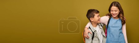 Smiling interracial preteen friends with backpacks hugging and having fun while celebrating international children day and standing on khaki background with copy space, banner 