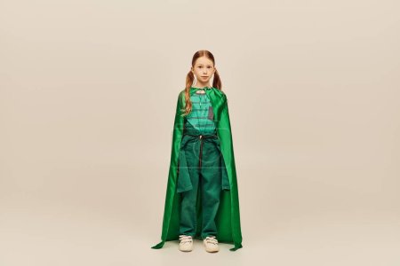 Redhead preteen girl in green superhero costume and cape looking at camera while standing on grey background during global child protection day celebration 