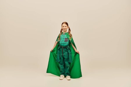Cheerful redhead kid in green superhero costume holding cape and looking at camera while standing on grey background in studio during international children`s day celebration