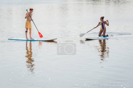 sportive african american woman in colorful swimsuit and young redhead man in swim shorts spending summer weekend while sailing on sup boards together