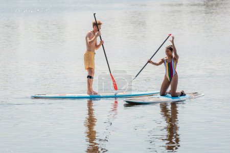 cheerful african american woman in colorful swimsuit kneeling on sup board near redhead man sailing with paddle while spending time on river in summer