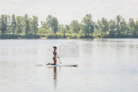 volle Länge der sportlichen afrikanisch-amerikanischen Frau im gestreiften Badeanzug, die auf Knien steht, während sie auf einem Surfbrett mit Paddel entlang des Flussufers mit grünen Bäumen segelt