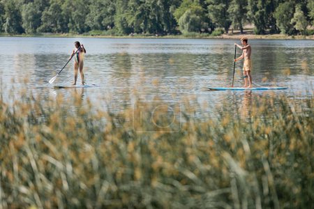aktywna wieloetniczna para w stroju kąpielowym spędzająca weekend na wodzie, żeglując po deskach surfingowych wzdłuż rzeki z zielonymi roślinami na niewyraźnym pierwszym planie w letni dzień