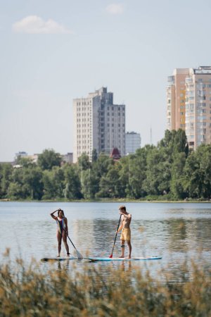 Sportliches Paar in Badebekleidung, das auf Surfbrettern mit Paddeln segelt, während es Zeit auf dem See mit malerischer Stadtlandschaft und Pflanzen im verschwommenen Vordergrund verbringt