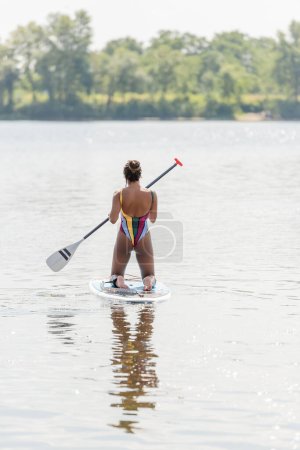 Rückenansicht einer aktiven afrikanisch-amerikanischen Frau im bunten Badeanzug, die auf Knien steht und das Paddel hält, während sie den Sommertag auf dem Sup Board auf dem See genießt
