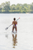 back view of active african american woman in colorful swimsuit standing on knees and holding paddle while enjoying summer day by sailing on sup board on lake puzzle #655804662