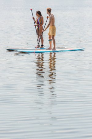full length of young redhead man in swim shorts and brunette african american woman in colorful swimsuit sailing on sup boards on calm river on summer day