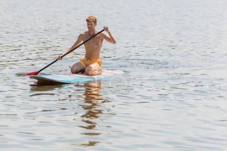 young and sportive redhead man in yellow swim shorts spending summer weekend on river while sitting on sup board and sailing with paddle during water recreation