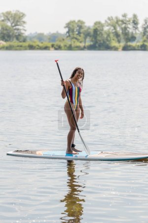 full length of overjoyed and active african american woman in striped swimsuit looking at camera while holding paddle and standing on sup board with picturesque riverside on background