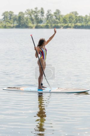 Foto de Longitud completa de la mujer afroamericana en traje de baño colorido de pie en sup board con paleta y mirando hacia otro lado mientras agita la mano en el lago en verano con pintoresca orilla del río en el fondo - Imagen libre de derechos