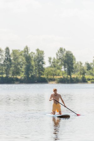 junger und aktiver Mann in gelben Badehosen mit Paddel und Knien auf dem Surfbrett, während er auf einem malerischen Fluss mit grünen Bäumen am Ufer mit malerischem Flussufer im Hintergrund segelt