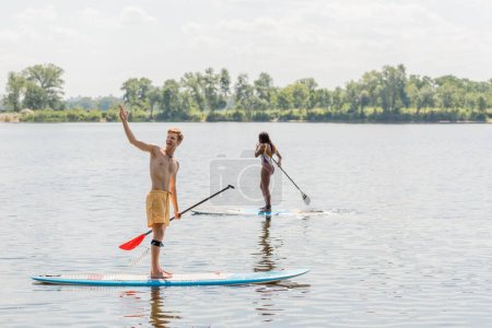 fröhlicher rothaariger Mann, der wegschaut und mit der Hand in die Nähe der sportlichen afrikanisch-amerikanischen Frau im bunten Badeanzug zeigt, die an einem Sommertag an Bord segelt, mit malerischem Flussufer im Hintergrund