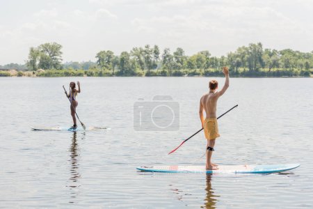 active interracial couple in colorful swimwear looking at each other and waving hands while sailing on sup boards on picturesque lake with green shore