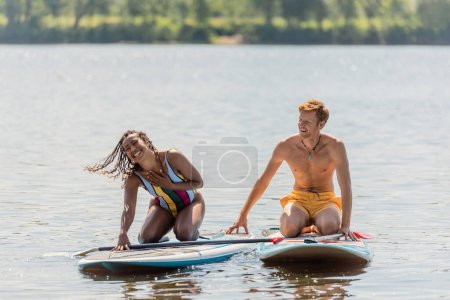Foto de Mujer afroamericana llena de alegría en traje de baño a rayas tocando el pecho y riendo cerca de hombre joven y pelirrojo mientras está sentado en tablas de sup en el lago en verano - Imagen libre de derechos