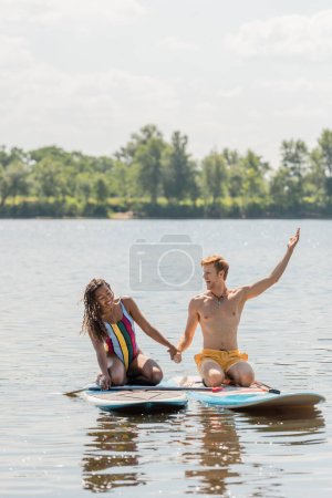 Unbekümmerter rothaariger Mann hält Hand in Hand mit fröhlicher afrikanisch-amerikanischer Frau und zeigt weg, während er auf Sup Brettern am See mit grünem malerischem Ufer sitzt