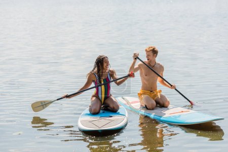 Unbekümmerte Afroamerikanerin und junger, fröhlicher Rotschopf in bunter Badebekleidung verbringen den Sommerurlaub am See und segeln auf Surfbrettern mit Paddeln