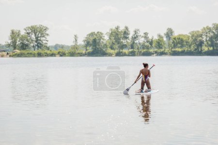 back view of active african american woman in striped swimsuit having rest on river by sailing on sup board near riverside with green trees, outdoor activity, summer vibes