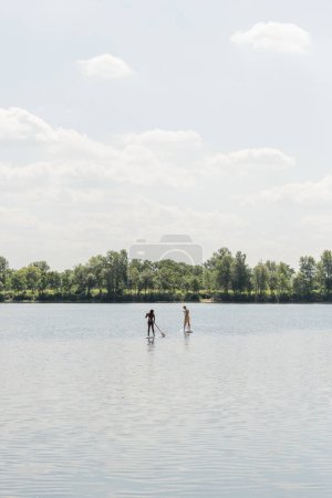 vista desde lejos de la pareja interracial deportiva navegando en tablas de sup mientras pasa el fin de semana en el lago con la orilla pintoresca verde bajo el cielo nublado, actividad al aire libre, vibraciones de verano