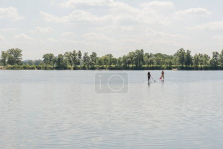 vista desde lejos de la pareja interracial y activa pasar el fin de semana de verano mientras navega en tablas de sup con paletas en el lago con la costa escénica bajo el cielo nublado