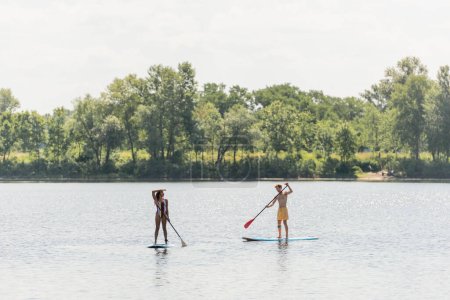 full length of african american woman sailing on sup board and looking away near young and sportive man in swim shorts on scenic lake with green trees on shore