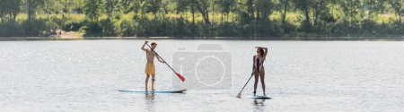 slender african american woman in colorful swimsuit looking away near young and sportive man sailing on sup board with paddle on river on summer day, banner