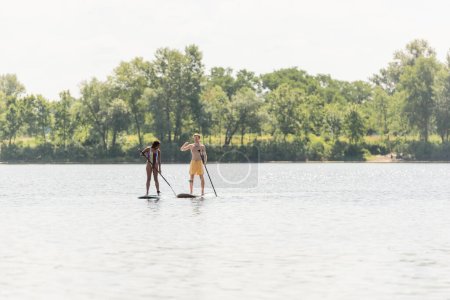 full length of sportive and active interracial couple spending time on lake while sailing on sup boards with paddles along shore with green summer trees