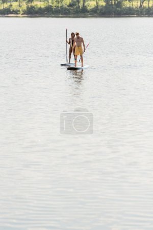 active multiethnic couple in colorful swimwear sailing on sup boards with paddles on calm river water near green bank during water recreation in summer