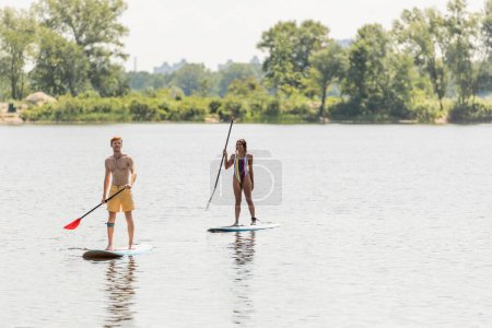 redhead man in swim shorts and african american woman in colorful swimsuit holding paddles while sailing on sup boards along green shore on summer day
