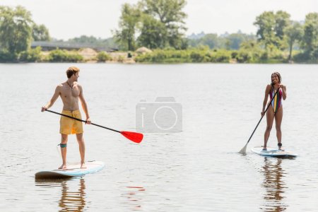 volle Länge des interrassischen sportlichen Paares mit Paddeln und lächelnd einander beim Segeln auf Surfbrettern auf dem Fluss während der Wassererholung am Sommerwochenende