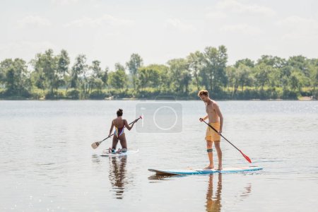 young and sportive redhead man standing on sup board with paddle near african american woman in colorful swimsuit sailing on picturesque lake with green shore