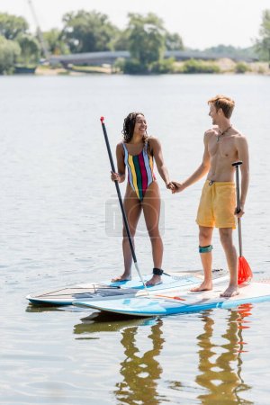 active redhead man and cheerful african american woman in striped swimsuit holding hands and smiling at each other while standing on sup boards on lake in summer