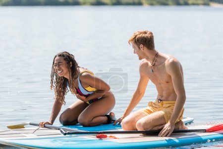 overjoyed african american woman in colorful swimsuit laughing near young and redhead man while having fun on sup boards during water recreation on summer weekend