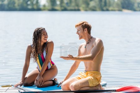 junger, rothaariger Mann, der mit der Hand zeigt und mit einer fröhlichen afrikanisch-amerikanischen Frau im bunten Badeanzug spricht, während sie zusammen auf Sup Brettern am See sitzt