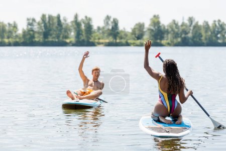 brunette african american woman in striped swimsuit and overjoyed redhead man waving hands at each other while sailing on sup boards on lake in summer
