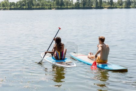 Téléchargez les photos : Vue de dos de la femme brune afro-américaine en maillot de bain rayé naviguant sur un sup board près de la rousse et l'homme sportif sur un lac pittoresque pendant la journée d'été - en image libre de droit
