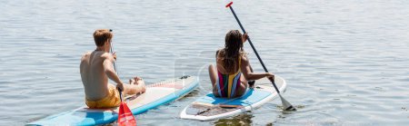 back view of young redhead man and sportive african american woman in striped swimsuit sitting on sup boards and sailing on lake on summer day, banner
