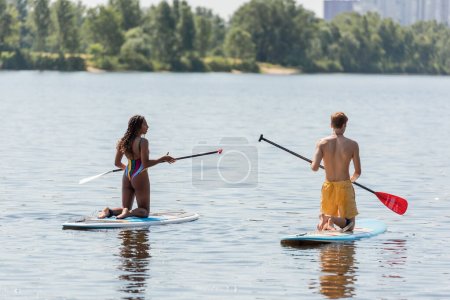 vue arrière de l'homme roux et de la femme afro-américaine en maillots de bain colorés debout sur les genoux sur des planches à soupe tout en tenant des pagaies et en naviguant sur un lac pittoresque