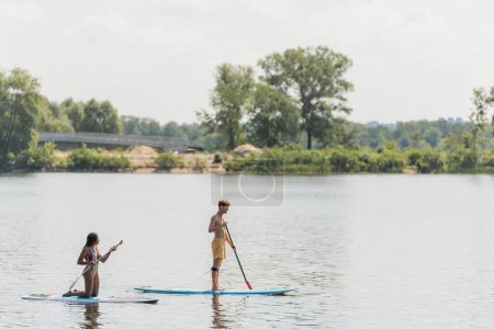 Photo for Side view of young sportive man and african american woman standing on knees and sailing on sup boards with paddles on river with green bank in summer - Royalty Free Image