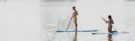 full length of redhead and sportive man sailing on sup board near african american woman in striped swimsuit on calm river during summer weekend, banner