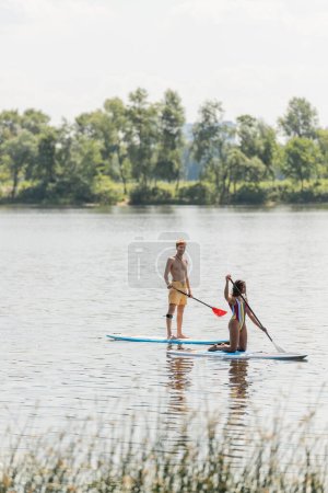 redhead and sportive man paddleboarding along green riverside near african american woman in colorful swimsuit during summer recreation on summer weekend